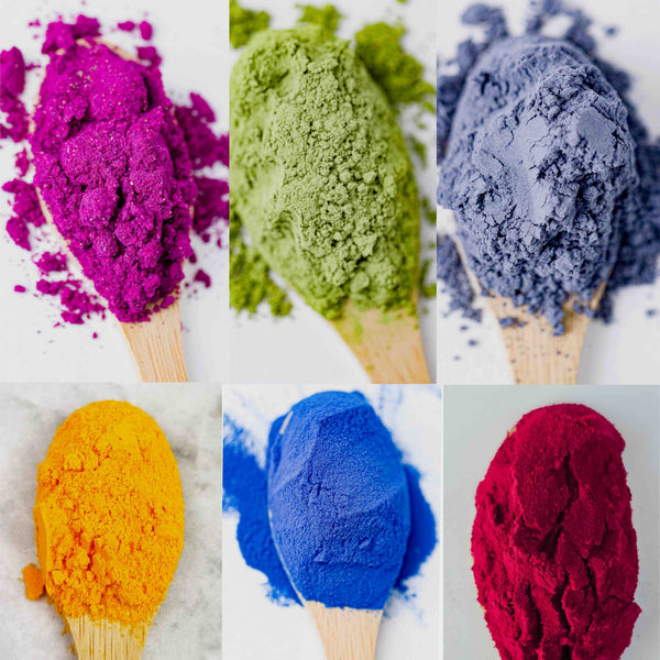 olorful botanical ingredient powders in purple, green, blue, yellow, bright blue and deep red on wooden spoons