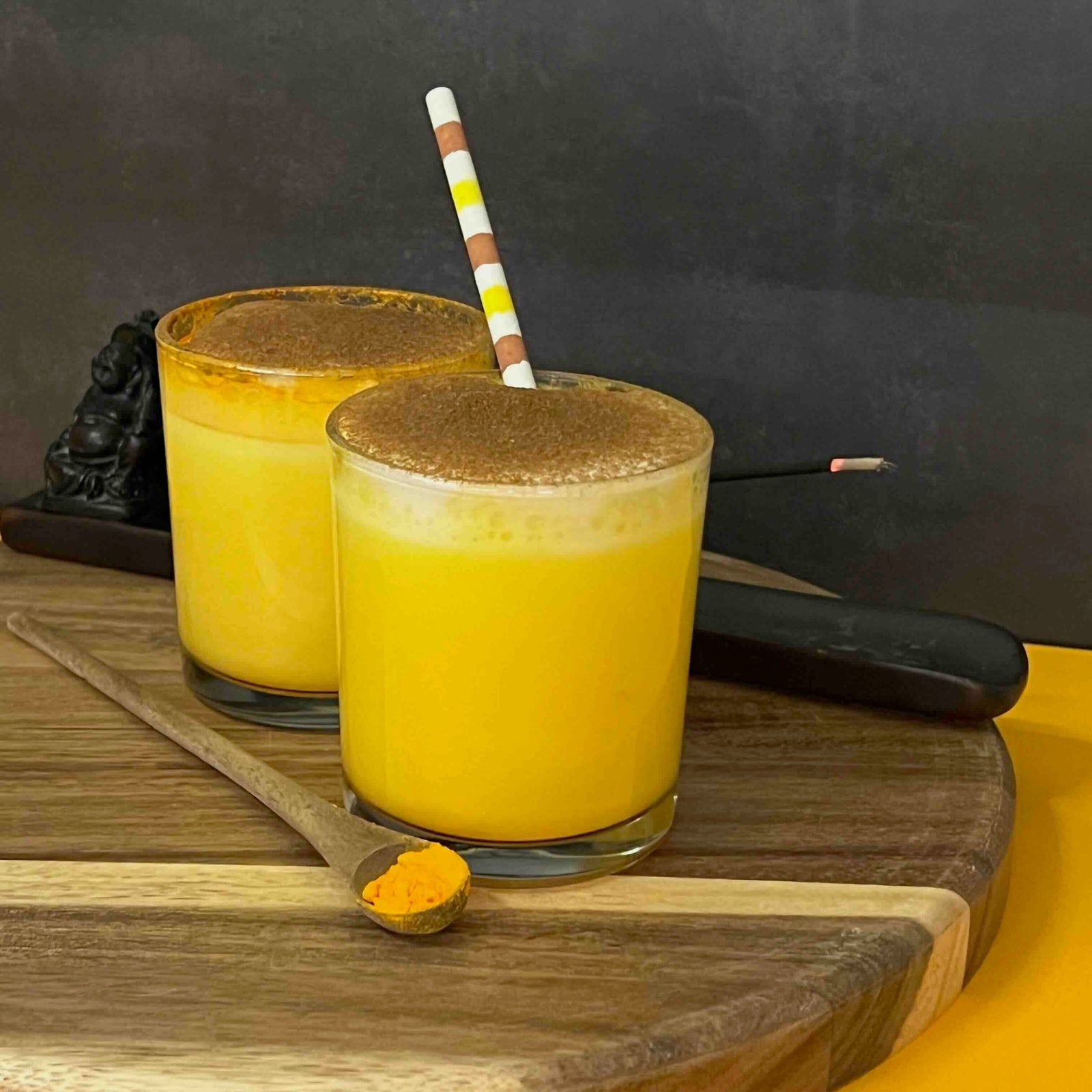 Two glasses of yellow smoothie with a brown topping on a wooden board, against a dark background.